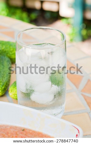 Shot of glass water with ice on the table. Part of plate with gazpacho and cucumbers.
