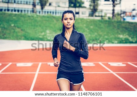 Indian woman running on track. Wearing long sleeve black top, black shorts with white stripe, pink runners. Long hair in pony tail. Looking into the lens. Multicultural, ethnic and inclusive theme.