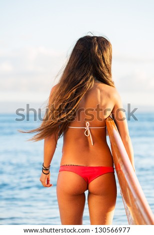 Rear view of beautiful sexy young woman surfer girl in bikini with surfboard on a beach at sunset