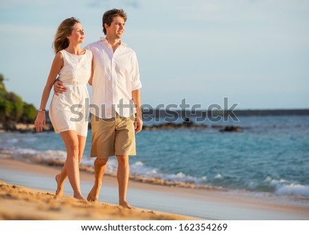 Romantic happy couple walking on beach at sunset. Smiling with arms around each other. Man and woman in love