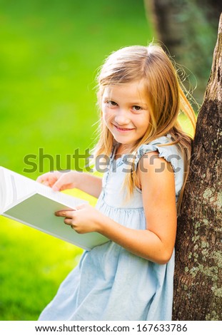 Cute little girl reading book outside on grass in backyard