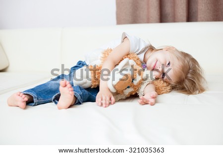 little blonde smiling girl three years sitting with a soft toy dog on a white leather couch