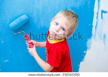 Cute blue-eyed blond pretty boy of five years in the red t-shirt makes repair paints the wall in blue roller
