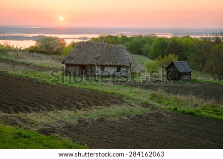 Agricultural land near old abandoned wooden house in Ukraine. Rural landscape with river on sunrise. Pathway to home.