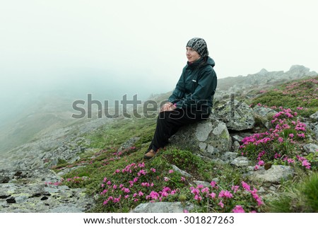 Woman in sport clothing sitting on the stone near blooming flowers in the rocky mountains. Fog covers the hill.
