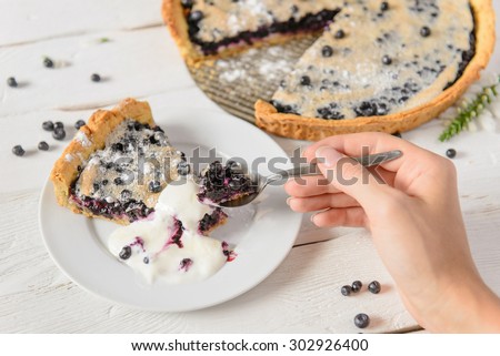 Eating blueberry pie on white rustic table served with cream by spoon in hand. Pile of berries near dessert.