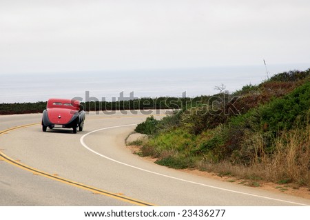 Photo of a classic car driving around a corner with the ocean in the back ground and the top down