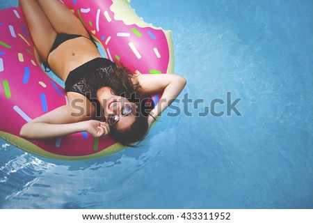 Young millennial girl in sprinkled doughnut float at pool, festival, hotel, beach, event smiling with sunglasses on during summer (with hipster matte texture)