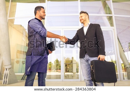 Handshaking between two young businessmen holding laptop - Concept of modern global market - Outdoor trade meeting between men of different race and culture - happy students shaking hands in the city