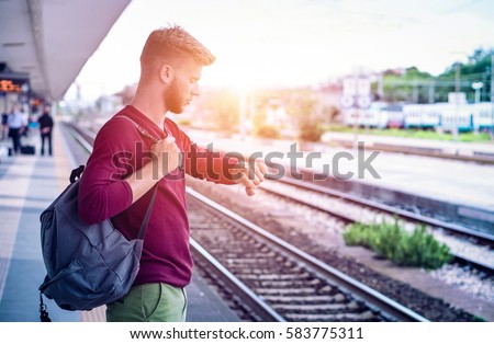 Young man checking the time on wristwatch at rail station platform - Student commuter waiting train at railway departures in pensive facial expression - Everyday lifestyle concept with sun halo filter