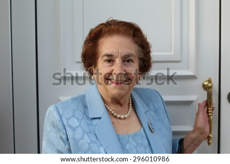 Smiling senior woman exiting her house pausing with her hand on the latch of the door to smile at the camera