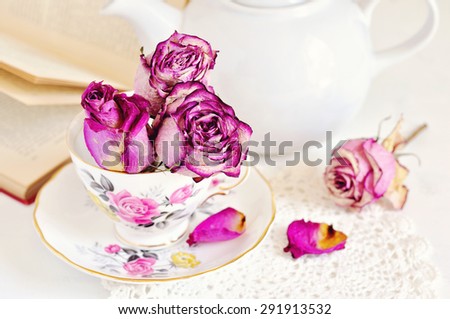 Vintage porcelain cup with dry roses and a vintage book on white background. Tea time table setting. Selective focus.