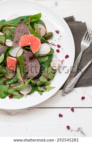 Salad with beetroot  leaves, grapefruit, duck breast, radish, capers and pomegranate seeds served with a slice of rye bread on a white plate on white wooden boards