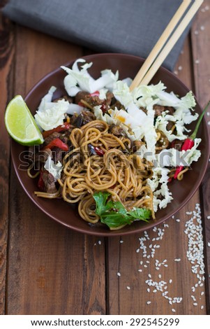 Chinese noodles cooked in a wok with beef and vegetables and served with a slice of lima, chinese cabbage and sesame seeds in a brown plate on wooden boards