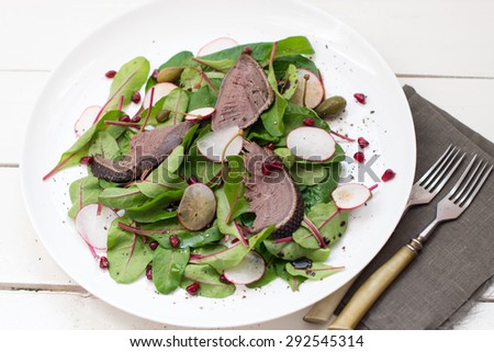 Salad with beetroot  leaves, duck breast, radish, capers and pomegranate seeds served with a slice of rye bread on a white plate on white wooden boards