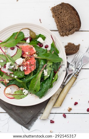 Salad with beetroot  leaves, grapefruit, feta cheese, radish, capers and pomegranate seeds served with a slice of rye bread on a white plate on white wooden boards