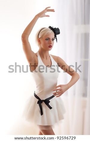 Beautiful young woman in white dress with small hat and black belt is acting as a living doll in a white empty room.