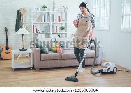 Smiling excited young housewife havig fun while cleaning floor with vacuum cleaner. Happy woman doing housework at home enjoy music wearing earphones. asian lady in arpon singing dancing house chores