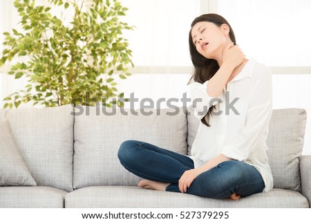 Young woman sitting on sofa with neck pain at home. lifestyle and health concept. mixed race asian chinese model.