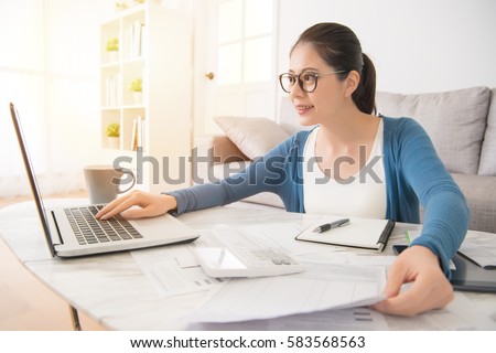 Happy young mixed race asian chinese woman calculating bills by laptop computer sitting on sofa in the living room at home. interior and domestic housework concept.