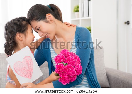 closeup photo of beautiful mother and cute daughter holding love card gift and pink carnation bouquet present hugs together on the sofa in the living room at mother\'s day.
