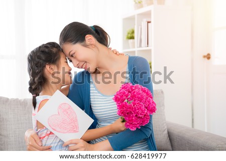 young asian mother accepting the little daughter hand painted card, embracing, little girl holding a pink flower girl, sitting on the sofa in the living room, happy looking at each other, copy space for mother\'s day.