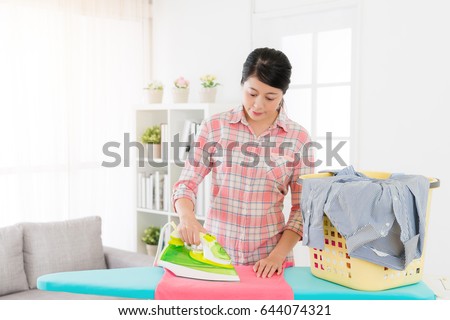 young pretty mother using iron on ironing board sort out messy clothing in living room. housewife doing housekeeping everyday at home concept.
