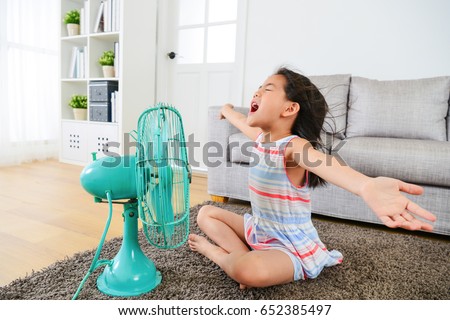pretty happy female kid opening arms enjoying cool wind from electric fan and sitting on floor in living room at summer vacation.