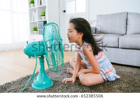 lovely youth little girl sitting on living room floor playing electric fan and enjoying cool wind in summer season at home.