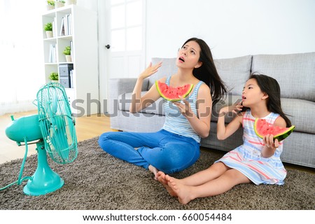 pretty mother with cute little daughter feeling very hot at summer day and eating watermelon blowing electric fan cool wind.