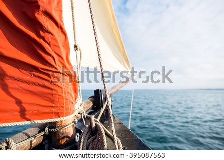 rope wound on a wooden cleat fixed on the hull of a rigging vintage sailing boat with a beige jib and an ocher sail filled by the wind at the blur background during a sunny sea trip in brittany