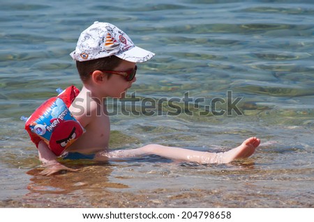 GREECE - JUNE 26: Boy with armbands, cap and sunglasses enjoying in the shallow sea water a sunny day on June 26,2014 on the beach in Nikiti, Sithonia peninsula, Greece.