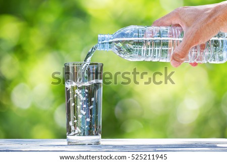 Man\'s hand holding plastic bottle water and pouring water into glass on wooden table on blurred green bokeh background
