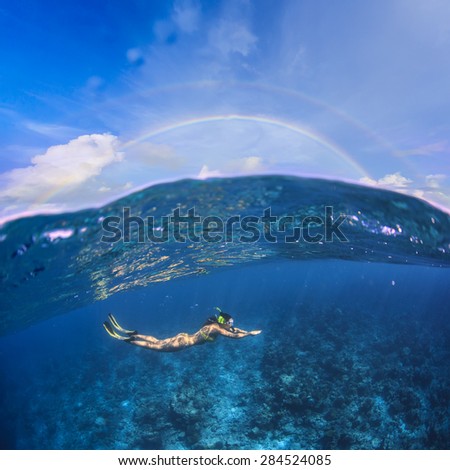 Underwater Sport Postcard. A female freediver with fins snorkeling under water surface in ocean. A rainbow appears on cloudy sky over beautiful seascape