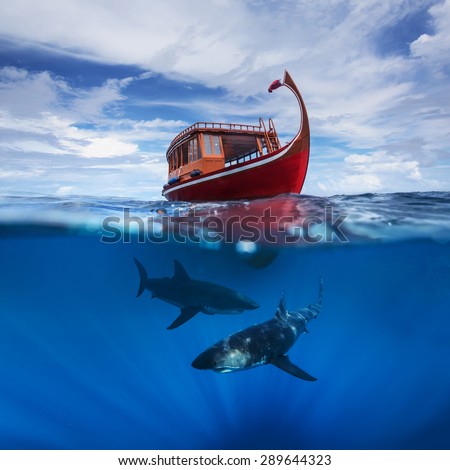 Half water ocean adventure. Two Great White Sharks hunting under red boat in deep blue. Maldivian ship floating on sea surface with mirrors of clouds on the tropical sky