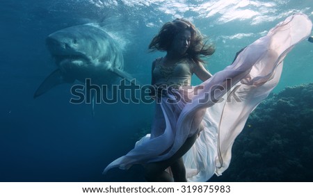 Most dangerous predator in The Ocean Great White Shark peaceful floating behind model posing underwater in fashion dress. Beautiful Model in deep sea in weightless condition hovering over coral reef.