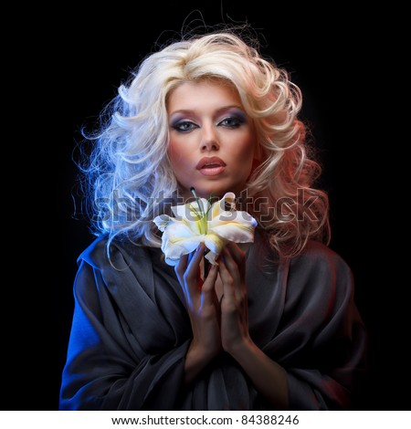 Attractive blonde with blue eyes wearing gray silk dress holding white lily by both hands on black background