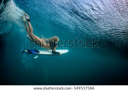 Blonde girl Surfer holding white surf board Diving Duckdive under Big Beautiful Ocean Wave. Turbulent tube with air bubbles and tracks after sea wave crashing. Ripples at water surface with blue color
