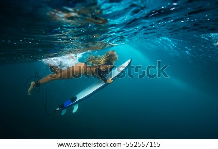 A blonde slim girl doing duck dive under blue wave. Water ripples under ocean surface. Water Sport activity in vacation. Sea fun lessons and beachlife lifestyle.