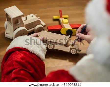 Santa Claus sitting in his workshop painting a toy airplane. Horizontal composition.