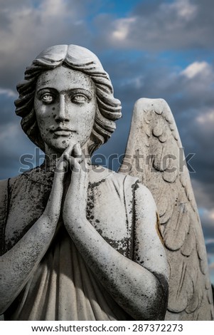 A statue of an Angel prays in front of a cloudy sky. The statue is weathered and corroded.