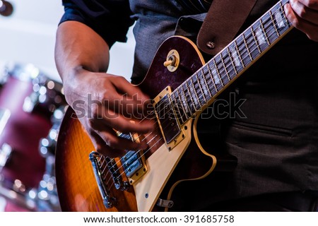 A hand strums at electric guitar strings in front of the blurred background of a drum kit as a lead guitarist plays a chord