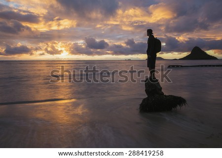 A person standing on a coconut tree stump in the shore line of Oahu\'s windward coast during a golden sunrise. The island known as China Man\'s Hat in the background.