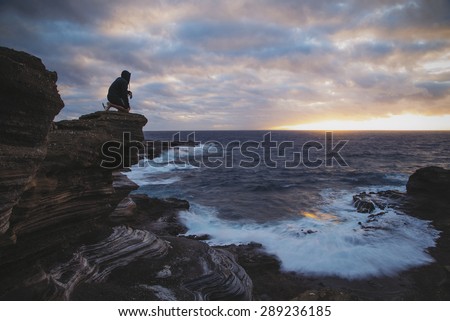 The silhouette of a man kneeling on a ledge looking out to sea as the sun peaks up over the horizon.