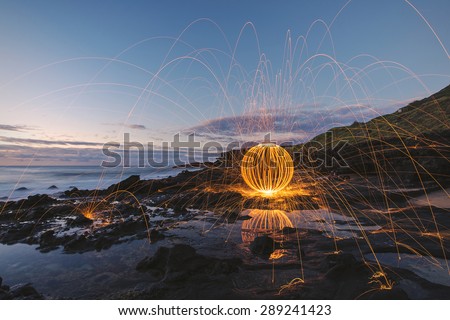 Long exposure of burning steel wool being spun into a sphere on the coastline before sunrise.