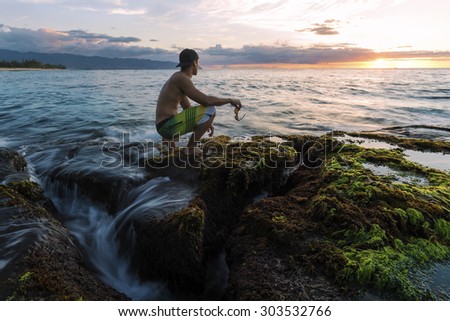 A man kneeling on a reef covered shore line holding glasses. The rocks covered in seaweed and flowing ocean water from the swells.