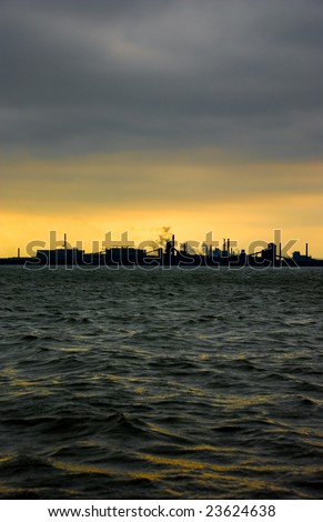 Silhouette of steel industry in Hamilton, Ontario, Canada against a yellow sky and dark clouds in front of water with waves reflecting yellow.