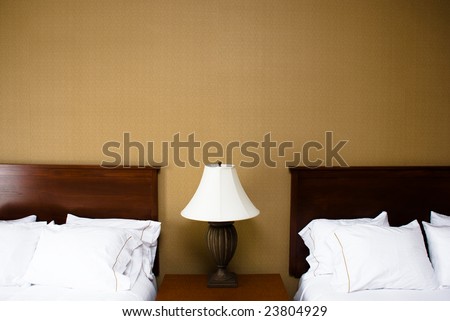 Two wood headboards and white fluffy pillows on either side of a lamp with white lampshade on a wooden bedside table top.  Beige plain wallpaper is on the wall behind the beds.