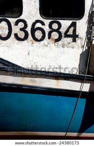 Part of a white, black and blue Maritime fishing boat.  Some of the windows show and a rope hangs down from out of frame.  Large black numbers are painted across the boat on peeling white paint.