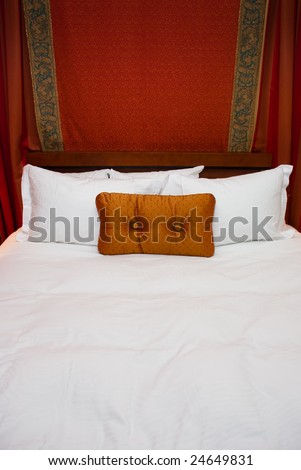 Orange pillow on big bed with white sheets.  Draped behind wood headboard is red fabric.